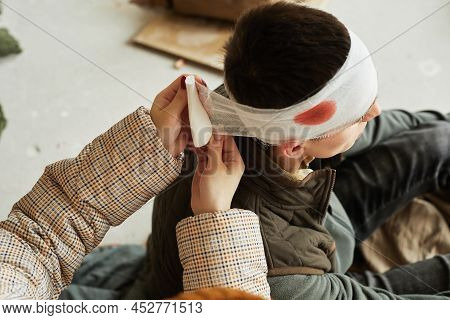High Angle Of Nurse Bandaging Head Of Wounded Civilian Man In Refugee Shelter, Copy Space