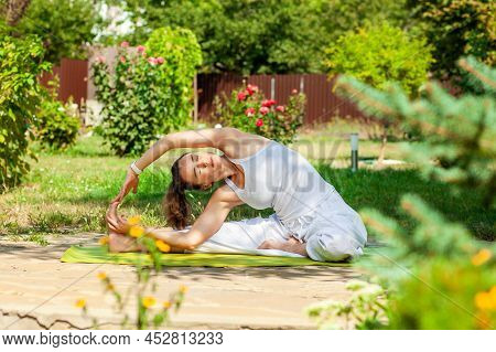Young Woman Practices Yoga In The Summer Garden - Inverted Tilt Of The Head To The Knee, Parivritta 