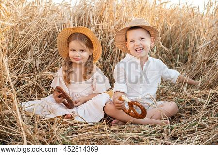 Happy Child Boy And Girl In Straw Hats Sitting In Wheat Field, Eating Bread On Summer. Cottage Core