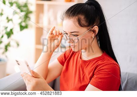 A Girl With Glasses With Vision Problems Is Trying To Read A Text On The Phone At Home. The Girl Has