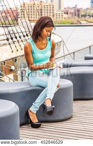 African American Woman Wearing Green Tank Top, Blue Fashionable Jeans, Sitting On Deck At Harbor, Cr