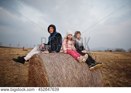 Three Kids Sitting On Haycock At Field.