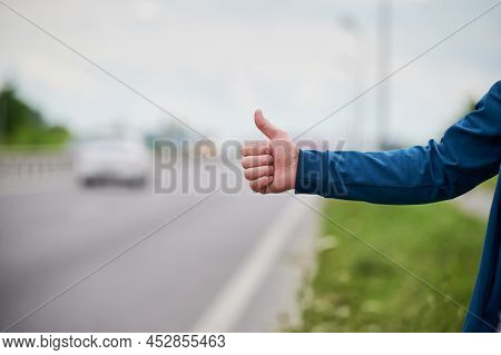 Close Up Of Mans Hand Hitchhiking By Roadside. Male Hand Showing Thumbs Up Gesture Outdoors On Blurr