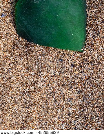 A Piece Of Glass On The Sand. A Shard Of Green Frosted Glass From A Bottle On A Seashell Beach.  A P