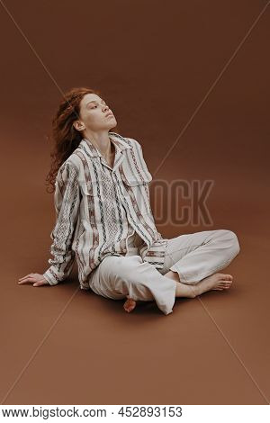 Portrait Of Young Woman With Red Hair Sitting In Relaxed Pose Against The Brown Background