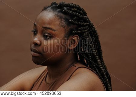Black Young Woman With Braids Sitting With Blank Expression Against The Brown Background
