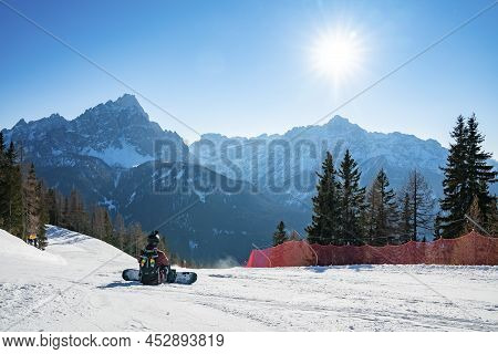 Sun Shining On Tired Snowboarder Sitting On Snowy Landscape Against Mountains
