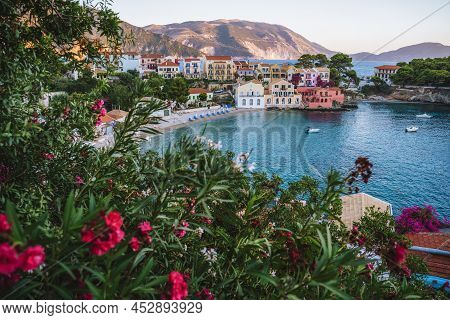 Assos Village In Evening Light On Kefalonia, Greece. Red Flower Blossom In Front Of Turquoise Bay An