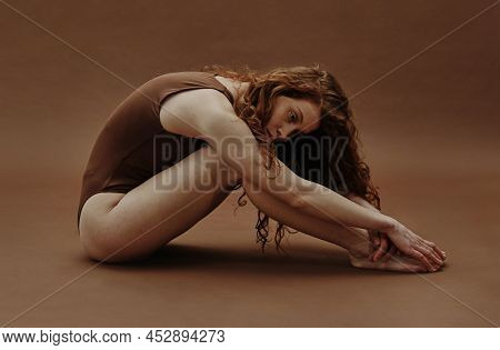 Young Pretty Woman With Red Hair Sitting In Swimsuit And Stretching Against The Brown Background