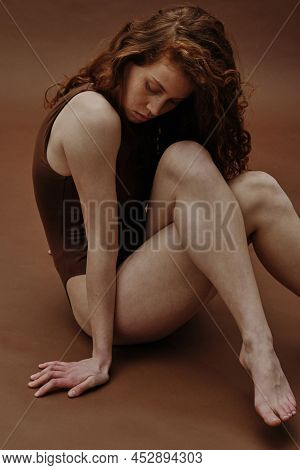 Young Ginger Woman With Slim Body Sitting In Swimsuit Against The Brown Background