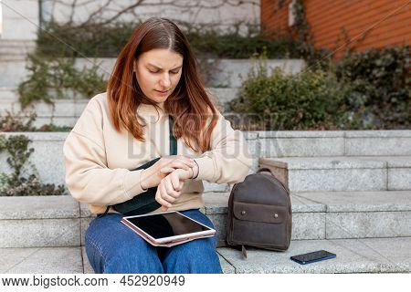 Woman Looking At Clock An Waiting Something. Cheerful Young Femile Sitting On Stairs On City Street,
