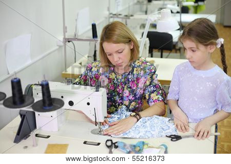 Female tailor sews at sewing machine and student girl looks at she. Focus on woman.