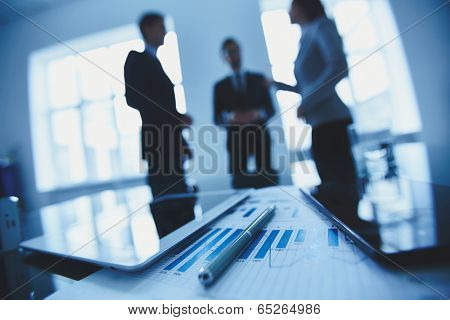 Close-up of business document, pen and touchpads at workplace on background of office workers interacting