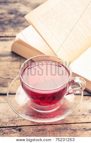 Cup Of Karkade Tea With Old Opened Book On Old Weathered Table