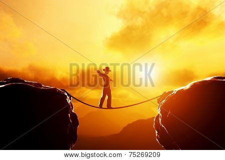 Man in hat walking and balancing on rope over precipice in mountains at sunset. Concept of business, risk taking, challenge, concentration.