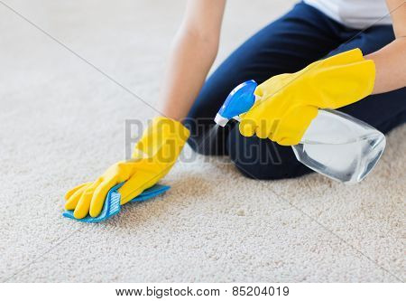 people, housework and housekeeping concept - close up of woman in rubber gloves with cloth and detergent spray cleaning carpet at home