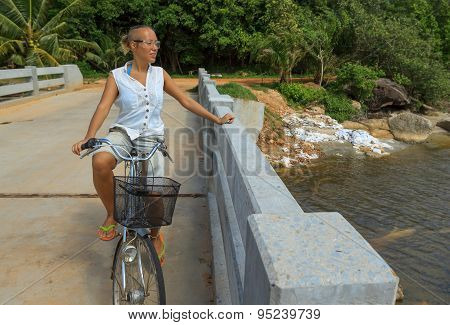 Young Woman Riding Bicycle Across River Bridge Next To Tropical Park