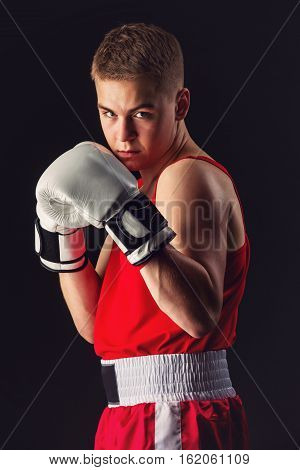 Young handsome boxer sportsman in red boxer suit and white gloves standing on black backgound. Copy space.