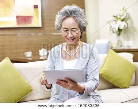 senior asian woman sitting on couch at home using a tablet computer.