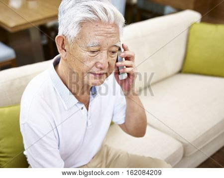 senior asian man sitting alone on sofa talking on mobile phone looking sad.