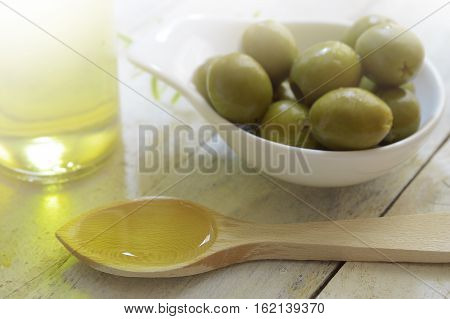 Spoon with olive oil next to an oilcan and some natural green olives. Scene on a wooden white table of a rustic kitchen.