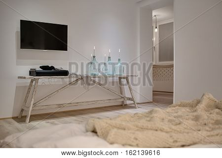 Modern interior with white walls and a parquet. There is a wooden table with candles in bottles, soundbar, shawl, TV, bed with a plaid, entrance to other room with hammock, glowing lamps. Horizontal.