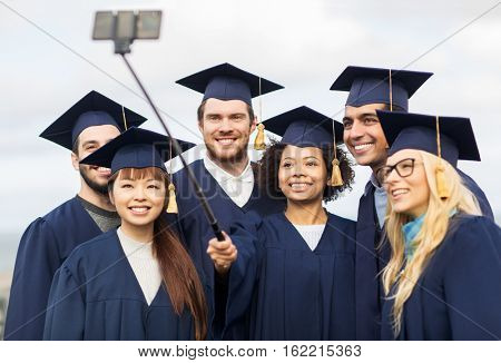 education, graduation, technology and people concept - group of happy international students in mortar boards and bachelor gowns taking selfie by smartphone outdoors