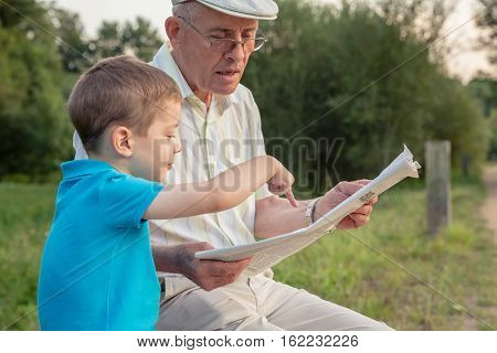Closeup of senior man reading newspaper and cute child pointing an article with his finger over a nature background. Two different generations concept.