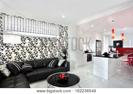 Modern living room interior designed by black vine which on the wall next to the kitchen. There are red lights hanging over the sink of the shiny black counter top next to the chair. The black sofa with pillows near the round black table