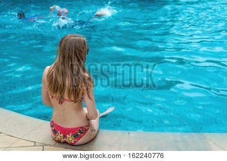 Back view of slim young beautiful woman in colorful swimsuit sitting on the edge of swimming pool. Enjoying tropical holidays