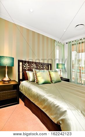 This modern bedroom of a luxury house decorated to get the gloss look for the whole room. The wooden bed is huge and there are caves pillows and bed sheets are shiny like tables lamps and the small metal cupboards also have a gloss look