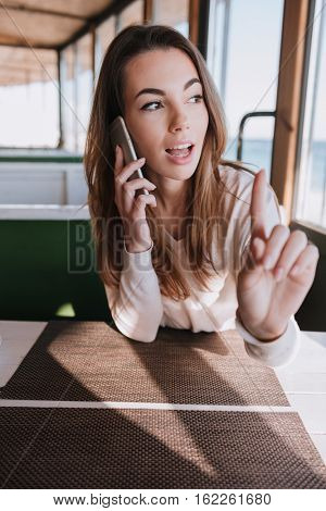 Vertical image of Woman sitting by the table and talking at phone on date in cafe near the sea