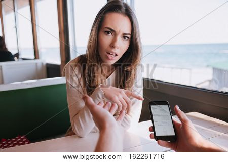Displeased woman sitting by the table on date in cafe near the sea with her man which using cell phone. First-person view