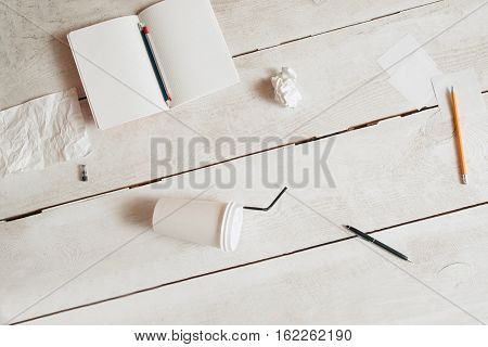 Workplace of untidy students flat lay, free space. Top view on blank copybook, pencils, coffee-to-go cup and paper wads scattered on white wooden table. Mess, disorder, bad form concept
