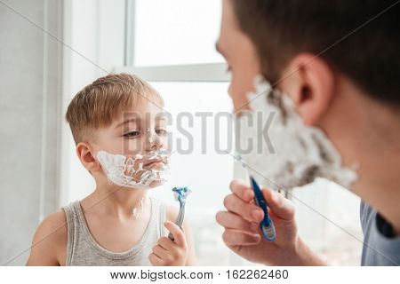 Image of funny father and son are applying shaving foam on their faces and smiling while shaving in bathroom