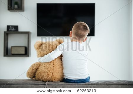 Back view image of cute little boy sitting on sofa with teddy bear waiting for parents at home and watching TV. Look at tv.