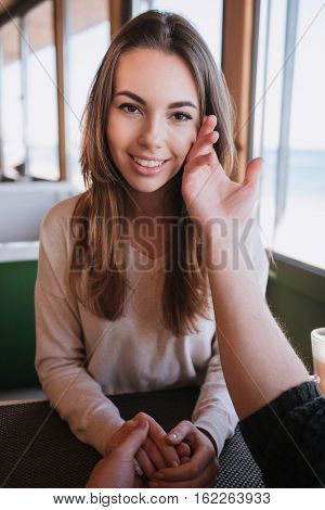 Vertical image of smiling woman sitting by the table on date in cafe near the sea and her man touching her cheek. First-person view