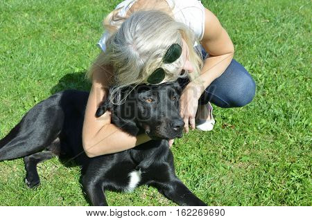 Woman hugging her adorable black dog who is lying on a green lawn
