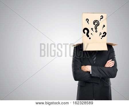 Businessman with folded arms and cardboard box with question marks covering head on grey background. Confusion concept