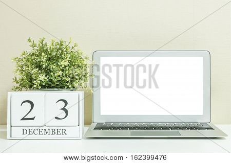 White wooden calendar with black 23 december word with white blank space at center of computer notebook on white wood desk and cream wallpaper textured background selective focus at the calendar