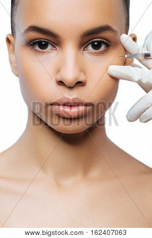 On the way of improvement. Pleasant involved young Negroid woman standing isolated in white background and expressing confidence while getting the injection in her face