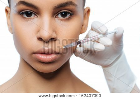 On the way of changes. Charming involved young Negroid woman standing against white background and expressing concentration while receiving the injection in her lips