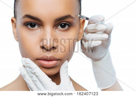 Forever young. Concentrated involved young Negroid woman standing against white background and expressing concentration while receiving the injection in her face