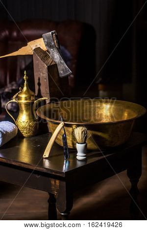 Vintage barber or shaver tools on wooden table. Old razor with cutthroat blade copper basin water jug axe shaving brush and towel in barbershop or hairdressing saloon