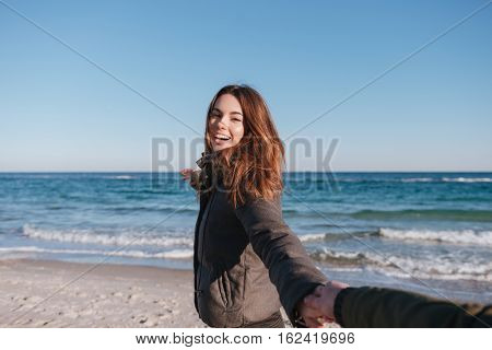 First person view of man holding hand of smiling woman in warm jacket on the beach