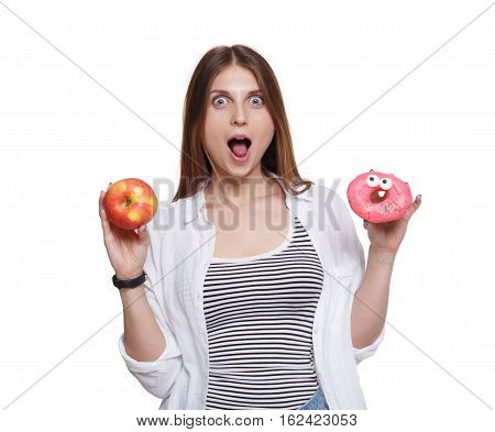 Choose healthy food, diet concept. Surprised long-haired young woman with big ripe red apple in one hand and a donut in another. Female portrait isolated on white background.