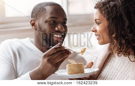 Full of romantic feelings. Cheerful careful playful African American couple sitting in the cafe and looking at each other while expressing joy and feeding each other with the cupcake
