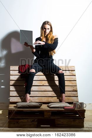 Pretty girl or beautiful woman cute sexy fashion model with long hair in casual clothes with laptop computer sits on backrest of wooden pallet sofa on white wall