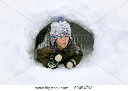 A cute young boy child is playing outside in a igloo fort tunnel he dug in a pile of snow on a winter day.