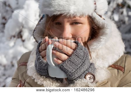 Woman In Grey Mittens, Coat And Hat With White Fur Drinking From Cup At The Background Of Winter For